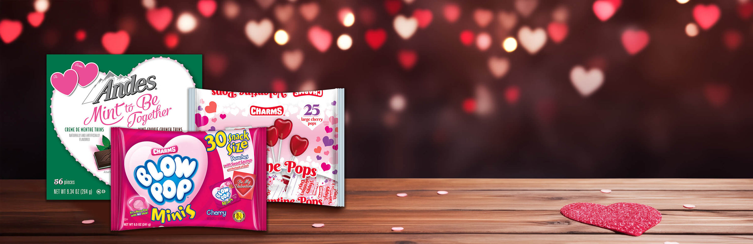 Valentine's candy display on a wooden table with a heart bokeh background. Includes Andes mints, Blow Pop Minis, and Charms Love Pops. Romantic and festive.